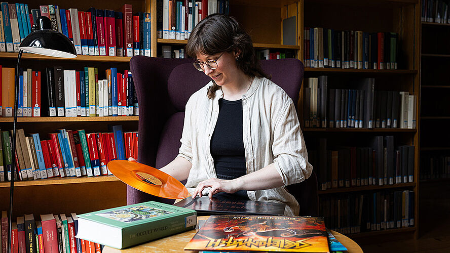 Querformatige Fotografie zeigt Lavinia Pflugfelder vor Bücherwand. Sie sitzt auf einem Sessel und häl Vinylplatten