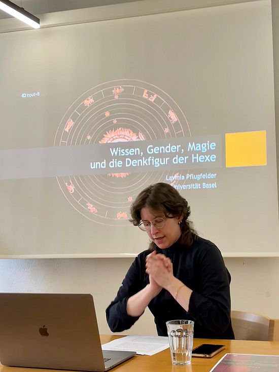 LAvinia Pflugfelder sitzt an einem Tisch, vor ihr ein Laptop, hinter ihr Projektion einer Powerpoint mit dem Titel "Wissen, Gender, Magie und die Denkfigur der Hexe".