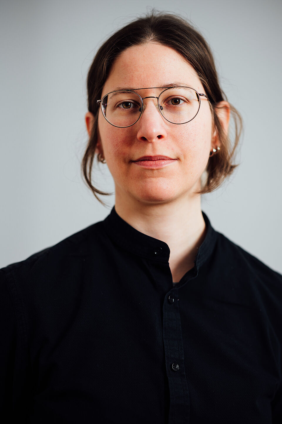 Portrait von Lavinia Pflugfelder: eine junge Frau mit Brille und halblangen dunkeln Haaren und schwarzem Hemd