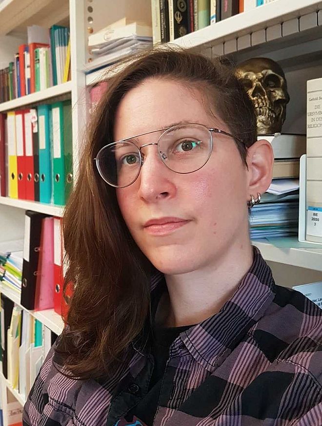 Portrait Lavinia Pflugfelder: junge weisse Frau mit schulterlangen braunen Haaren und undercut, trägt eine Brille und lächelt, Bücherregal im Hintergrund.